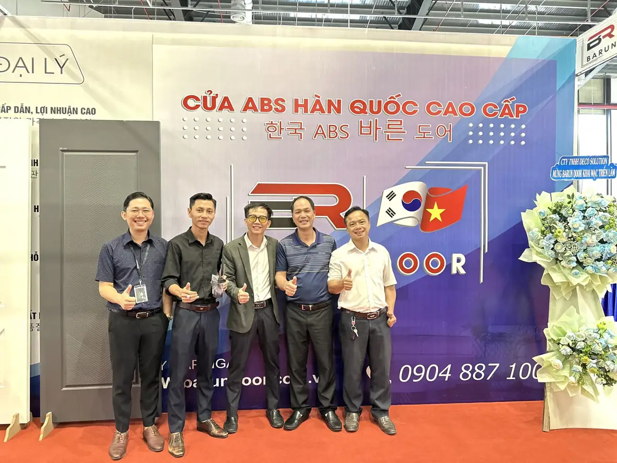Nhóm doanh nhân chụp ảnh lưu niệm trước phông gian hàng Barun Door bên cạnh mẫu cửa xám và kệ hoa